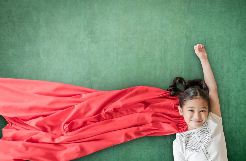A young girl laying on a green surface with a red cape striking a superhero pose symbolizing empowerment