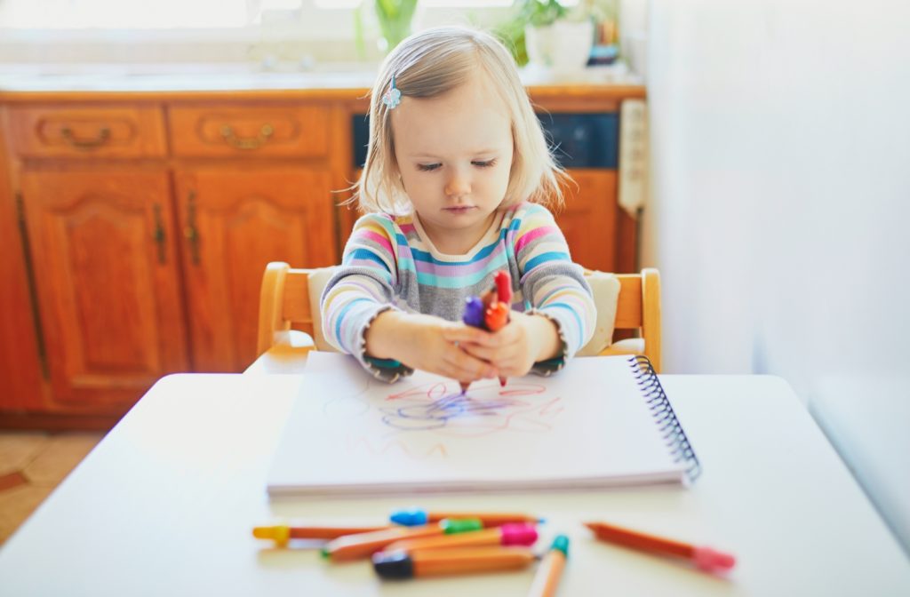 Young blonde girl holding four markers with both hands concentrating on her workbook.
