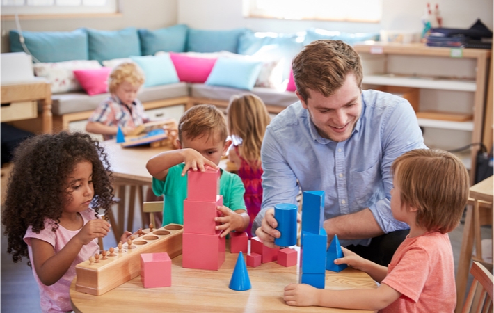 A young boy plays with a Montessori pink tower at a table with two other children, while an educator is helping one of them.