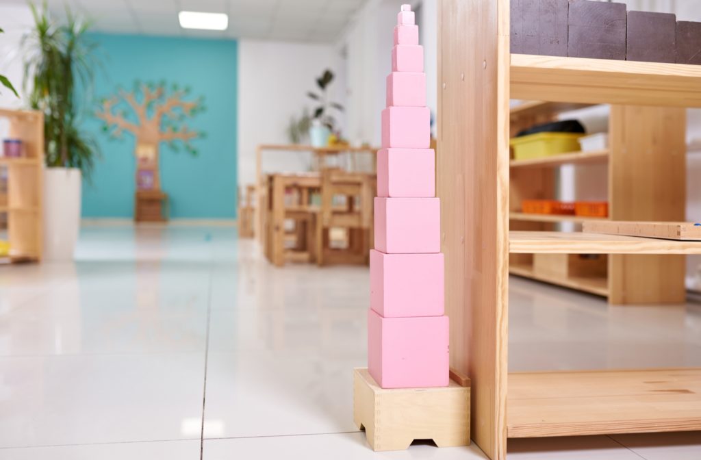 A Montessori pink tower stacked up next to an empty bookshelf inside a brightly lit classroom.