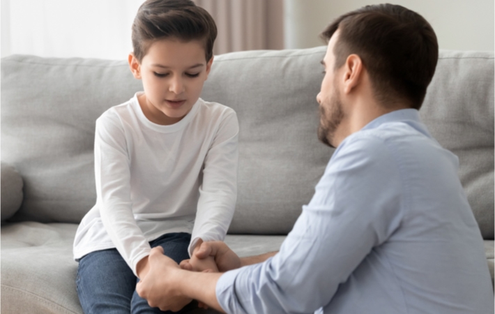 Young brunette boy sitting on a grey couch holding his dad's hands as his dad kneels in front of him to talk to him.