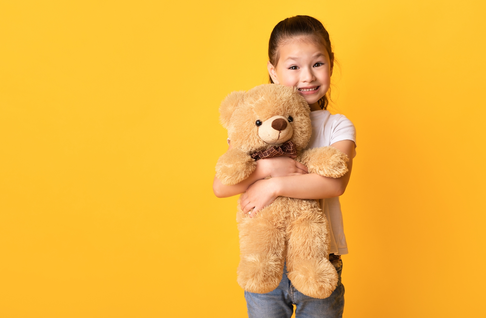 Young smiling girl tightly hugs a large teddy bear as she stands in front of a sunset-yellow background.