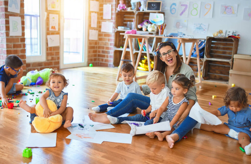 A female teacher is sitting on the floor with a group of toddlers while they are colouring their did some art.