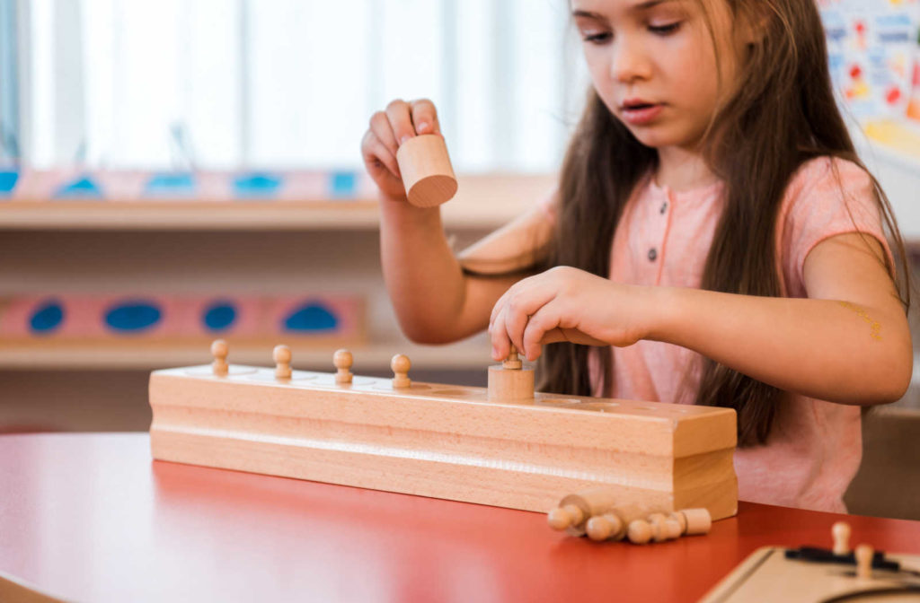 A young child sitting at a wooden table, focused on a Montessori educational game in front of them. The game consists of a series of colored wooden blocks, arranged in a specific pattern. The child is using their hands to manipulate the blocks and solve the puzzle.