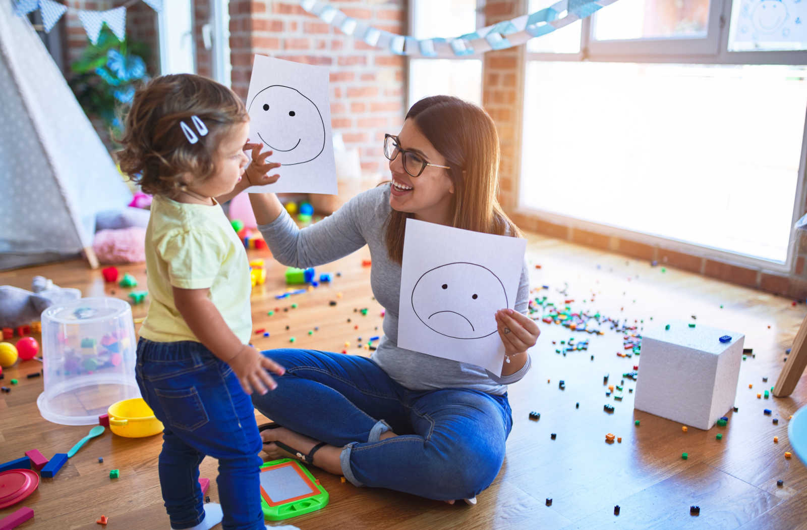 A female teacher is sitting on a floor and holding emoticons with her hands teaching a toddler a sad and a happy emotion.