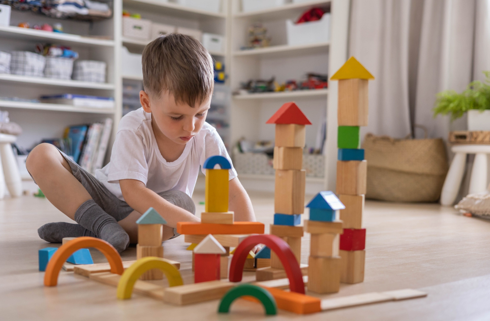 A young boy in a Montessori school, sitting on the floor and playing with wooden toy bricks.