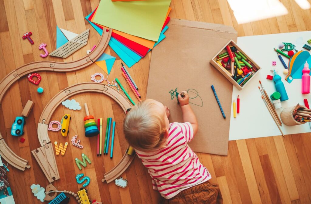 A young child on the floor draws on cardboard with art supplies to support hands-on learning, surrounded by colourful Montessori materials.