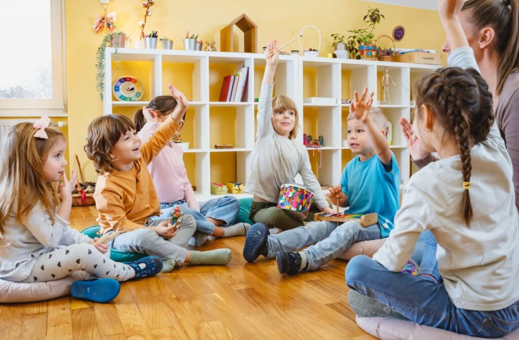 6 students and a teacher gather in a circle on the wooden floor of a Montessori classroom. Four children have their arms raised, ready to contribute to the conversation.