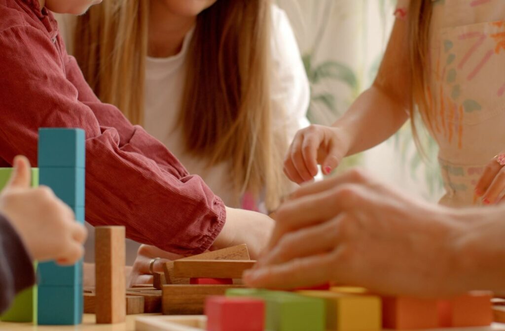 a close-up image of a group of young students using their hands to play with building blocks together. A teacher with long hair is supervising in the background.