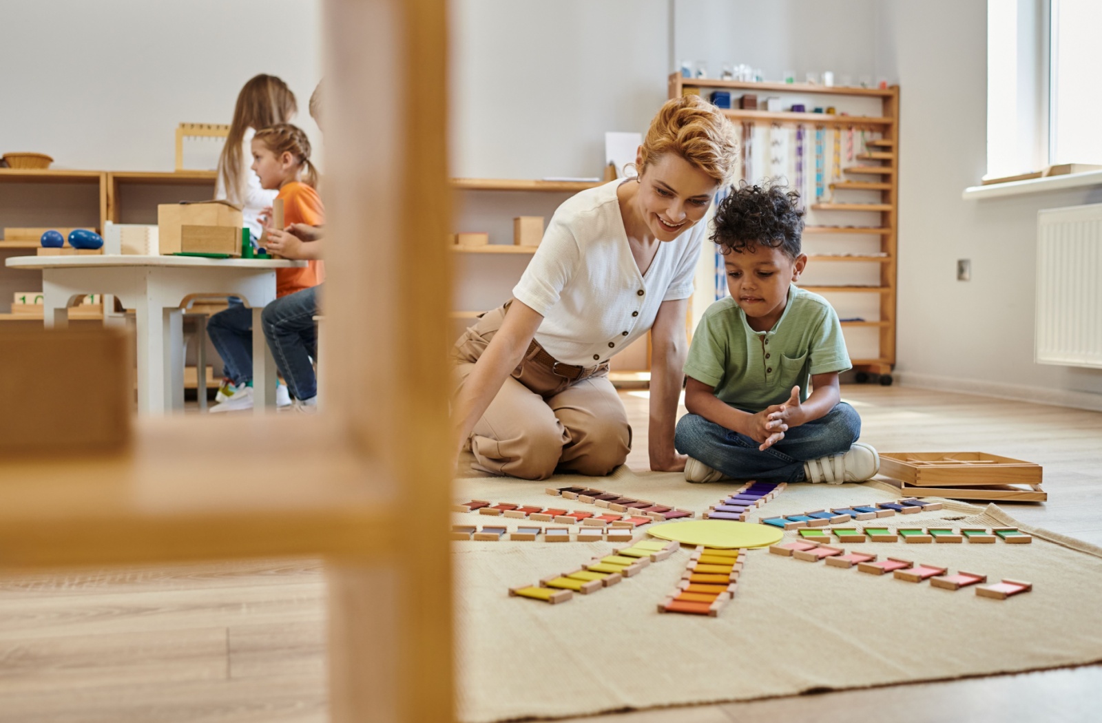 A teacher sitting on the floor with a child as they play and learn in a Montessori classroom.