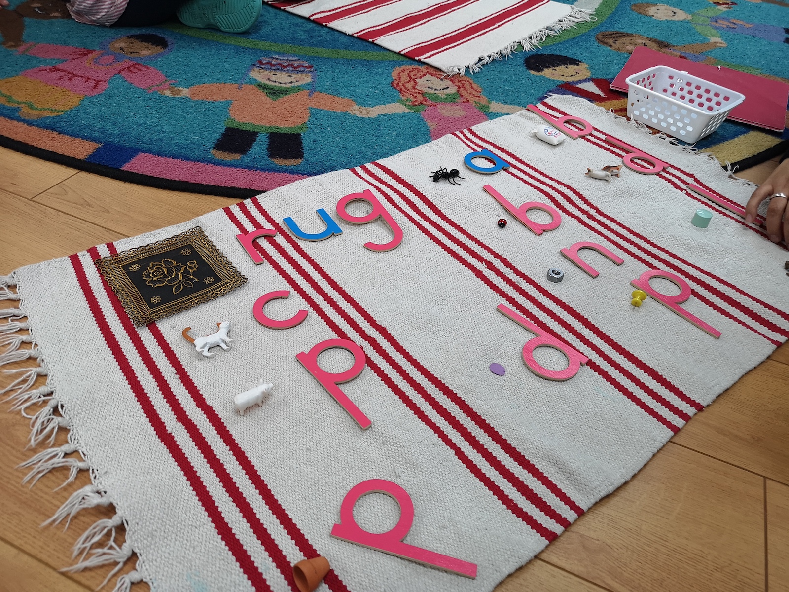 A Montessori rug with red and white stripes, displaying pink and blue tactile letters alongside small objects that start with the corresponding sounds, such as animals, buttons, and miniature items, used for phonetic learning.