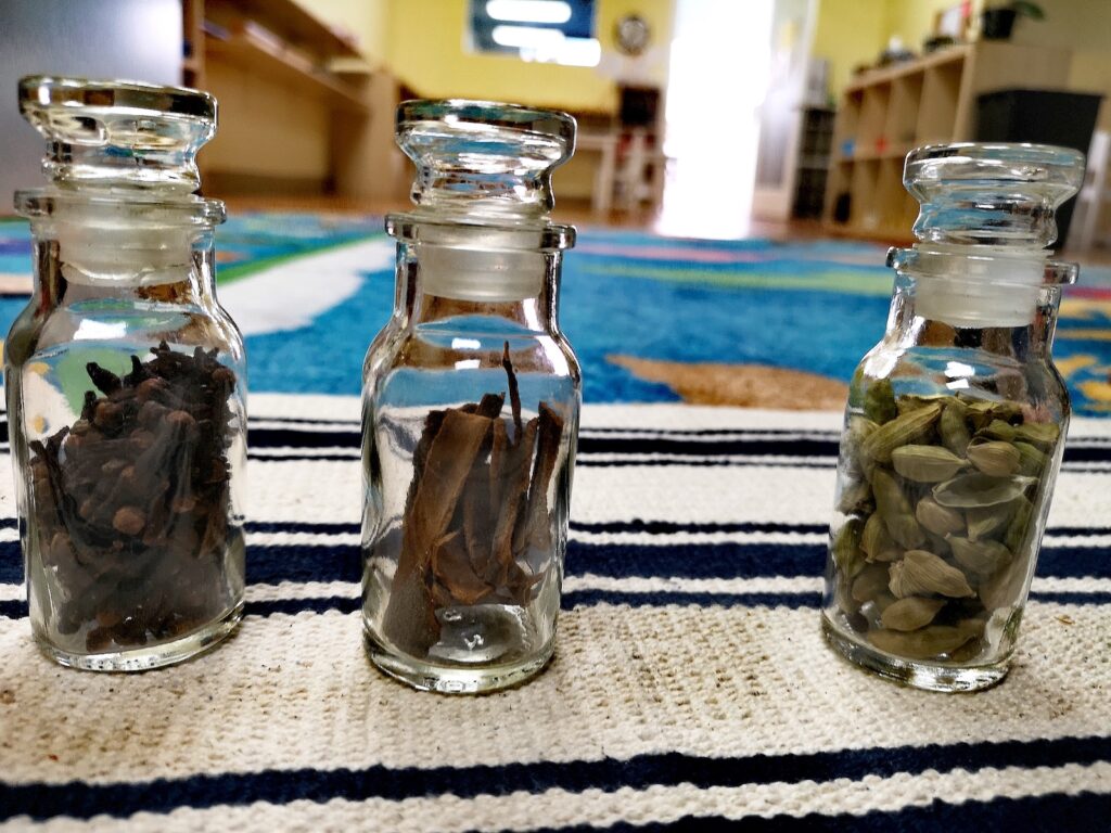Three small glass jars with stoppers containing different spices – cloves, cinnamon sticks, and green cardamom pods – arranged on a striped mat in a Montessori classroom.