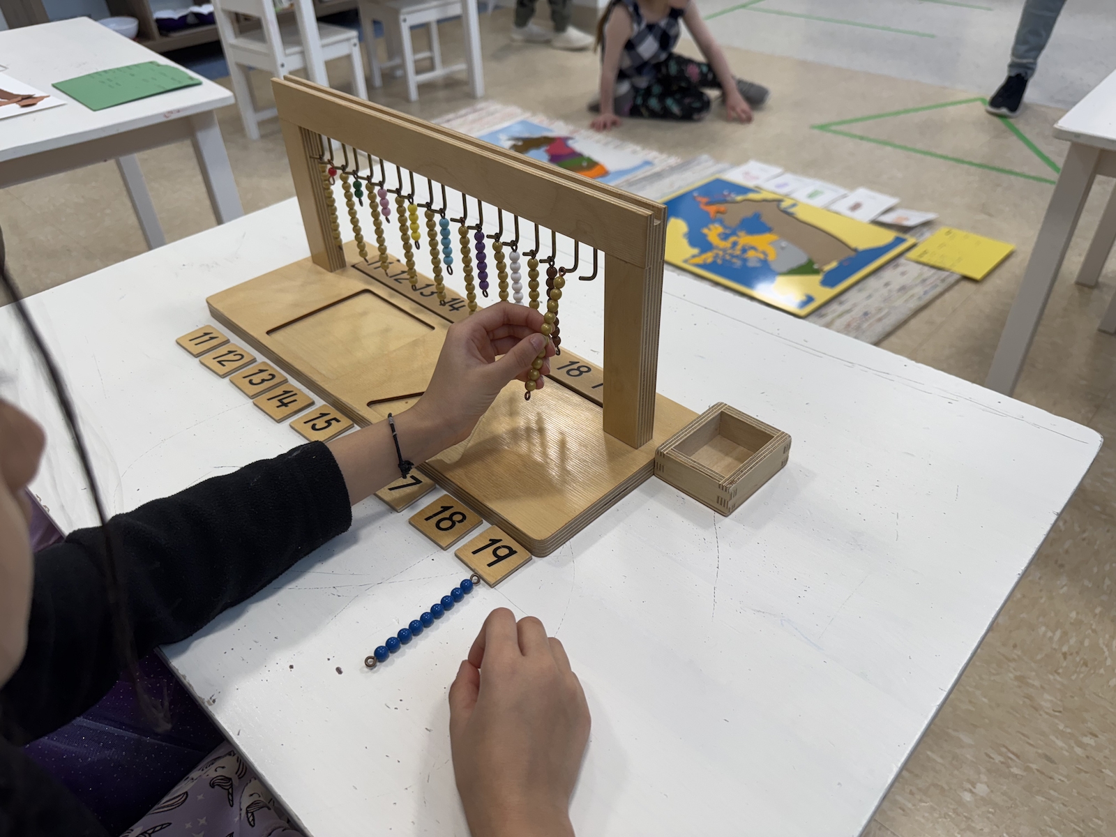 Child working with a Montessori math bead frame on a white table. The child is counting and moving yellow beads while number tiles from 11 to 19 are placed below the frame. In the background, other children are engaged with Montessori materials, including a large map.