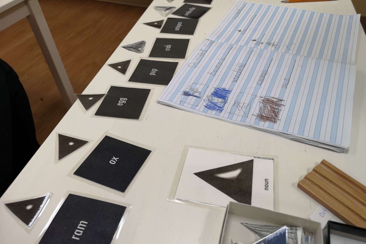 A collection of Montessori grammar learning materials on a white desk, featuring black square cards with words like 'ram', 'ox', and 'egg', each accompanied by a small black triangle symbol representing a noun. Beside these, an open notebook contains a child's drawings and handwriting, and a larger card labeled 'noun' shows a prominent black triangle.