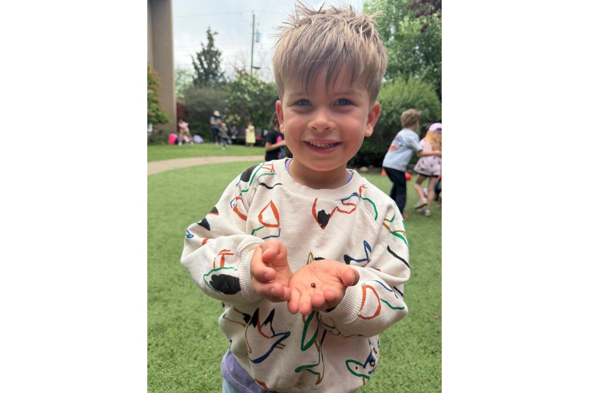 A smiling young child stands outdoors on a grassy play area, proudly holding a tiny ladybug in open hands and showing it to the camera. The child has tousled light brown hair and wears a cream-colored sweatshirt with colorful abstract line drawings. Other children play in the softly blurred background among trees and bushes on an overcast day.